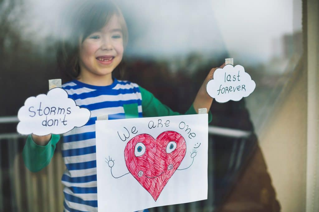 A boy stands at the window, holding up a painting of a heart with a smile to remind neighbors that we are all in this together and it won’t last forever.