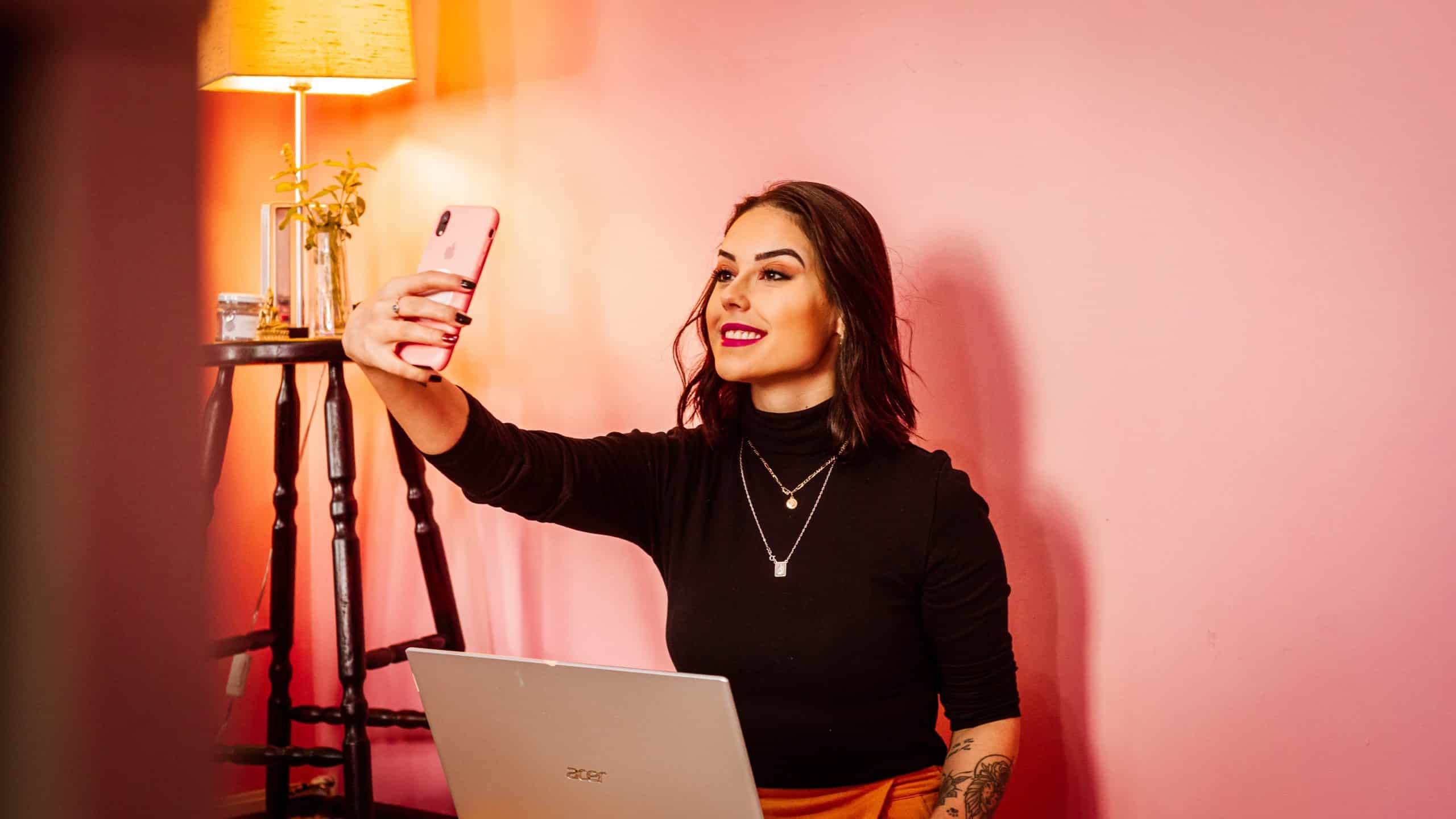  A woman with her laptop on her computer takes a selfie with a pink wall in the background creating user-generated content.