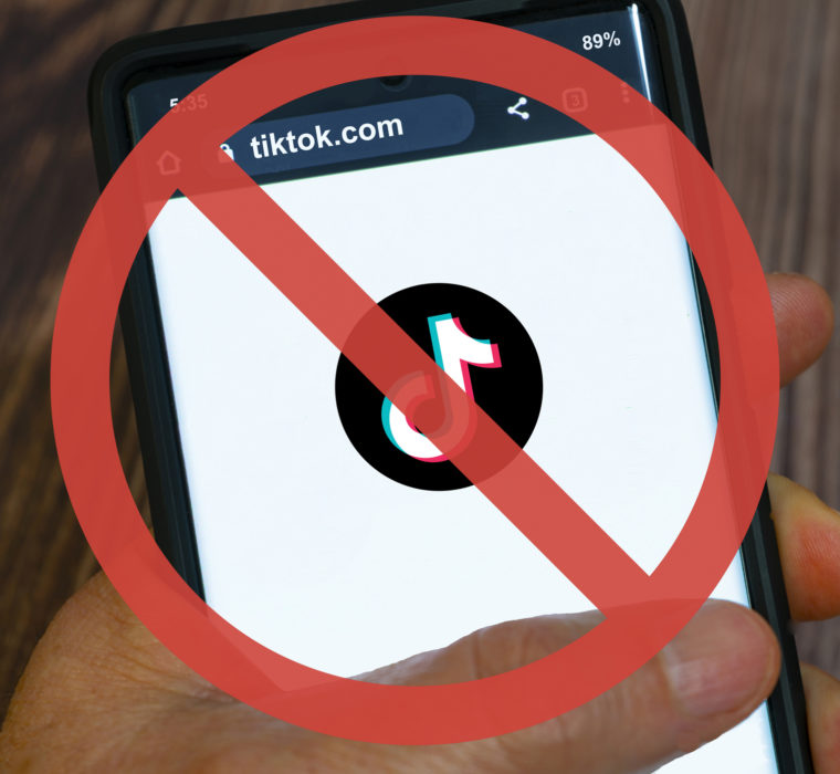 An image of someone holding a smartphone with the TikTok logo on it and a vector image over the top of the photo of a circle crossed out over the phone to represent TikTok ban.