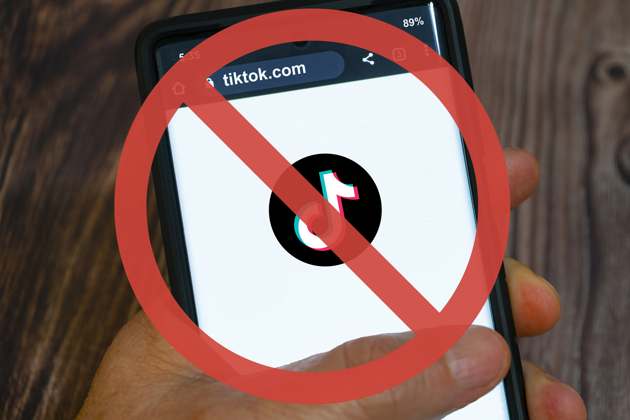 An image of someone holding a smartphone with the TikTok logo on it and a vector image over the top of the photo of a circle crossed out over the phone to represent TikTok ban.